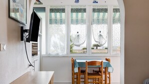 A room with a wooden table, chairs, a TV mounted on the wall, and a window with patterned curtains. - Studio Apartment "Monolocale 12" with Sea View, Wi-Fi & Air Conditioning (Pietra Ligure)