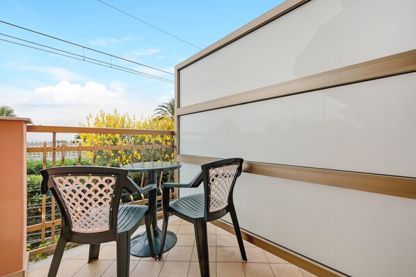 A balcony with two chairs and a table, a white wall, and a view of yellow flowers and a fence.