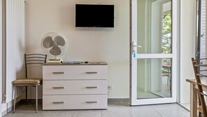 Une pièce avec un téléviseur fixé au mur, un ventilateur posé sur une commode, une chaise et une porte-fenêtre coulissante donnant sur un espace extérieur.