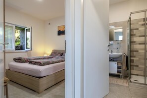 Ein Schlafzimmer mit Bett, Nachttisch, Fenster mit Vorhängen, Bad mit Waschbecken, Spiegel und Dusche.
