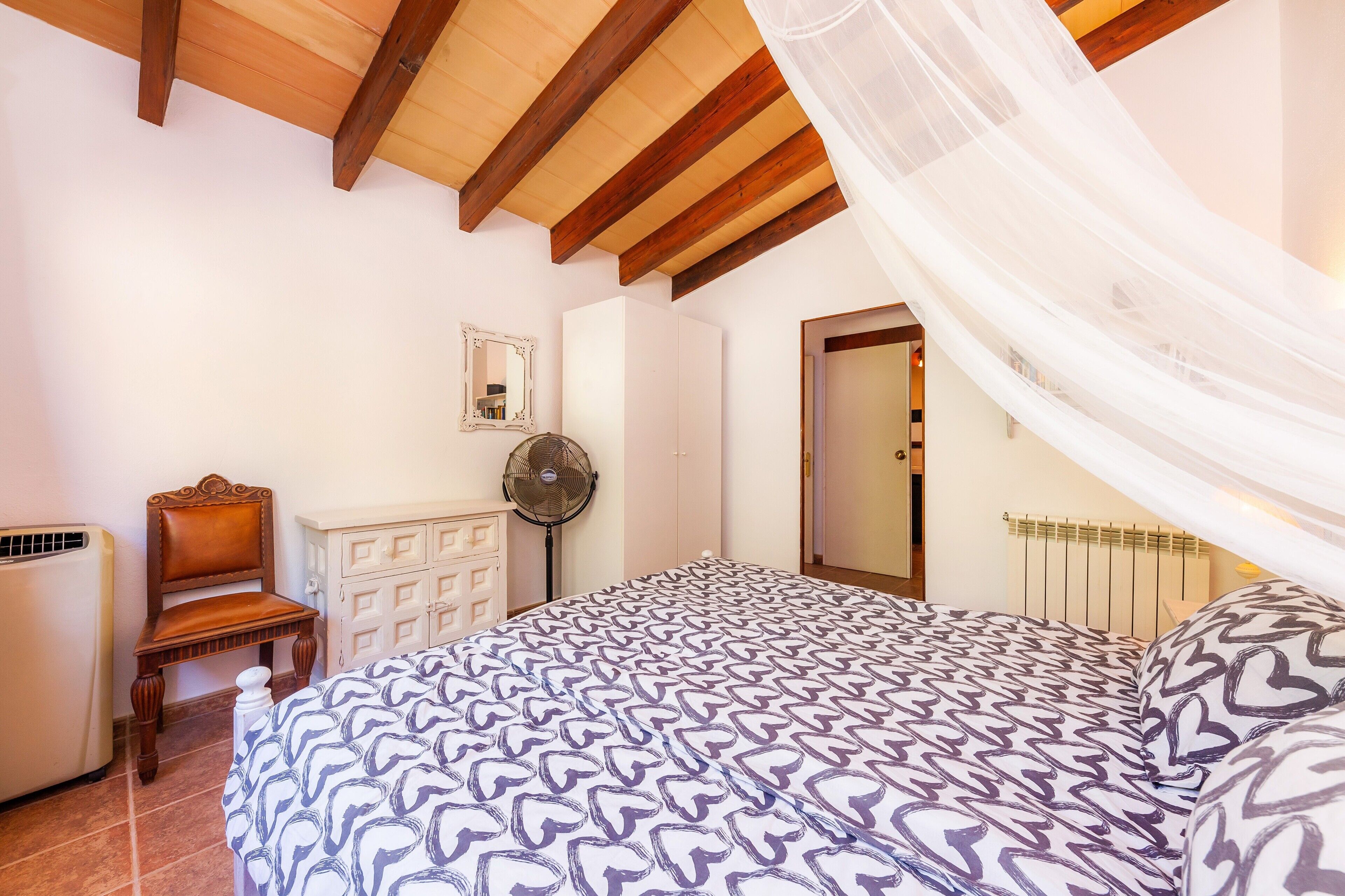 A bedroom with a bed, a chair, a fan, a radiator, and a door.