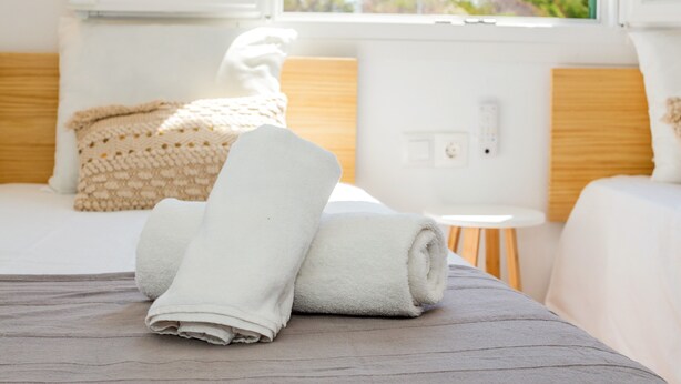 A neatly made bed with a rolled white towel, a small round table, and a window with a view of greenery. - Apartment 'Delfín 8' with Shared Pool, Wi-Fi, and Air Conditioning (Ciutadella de Menorca)