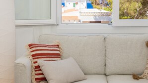 A beige sofa with two pillows, a window with a view of buildings and trees, and a white wall. - Apartment 'Delfín 8' with Shared Pool, Wi-Fi, and Air Conditioning (Ciutadella de Menorca)