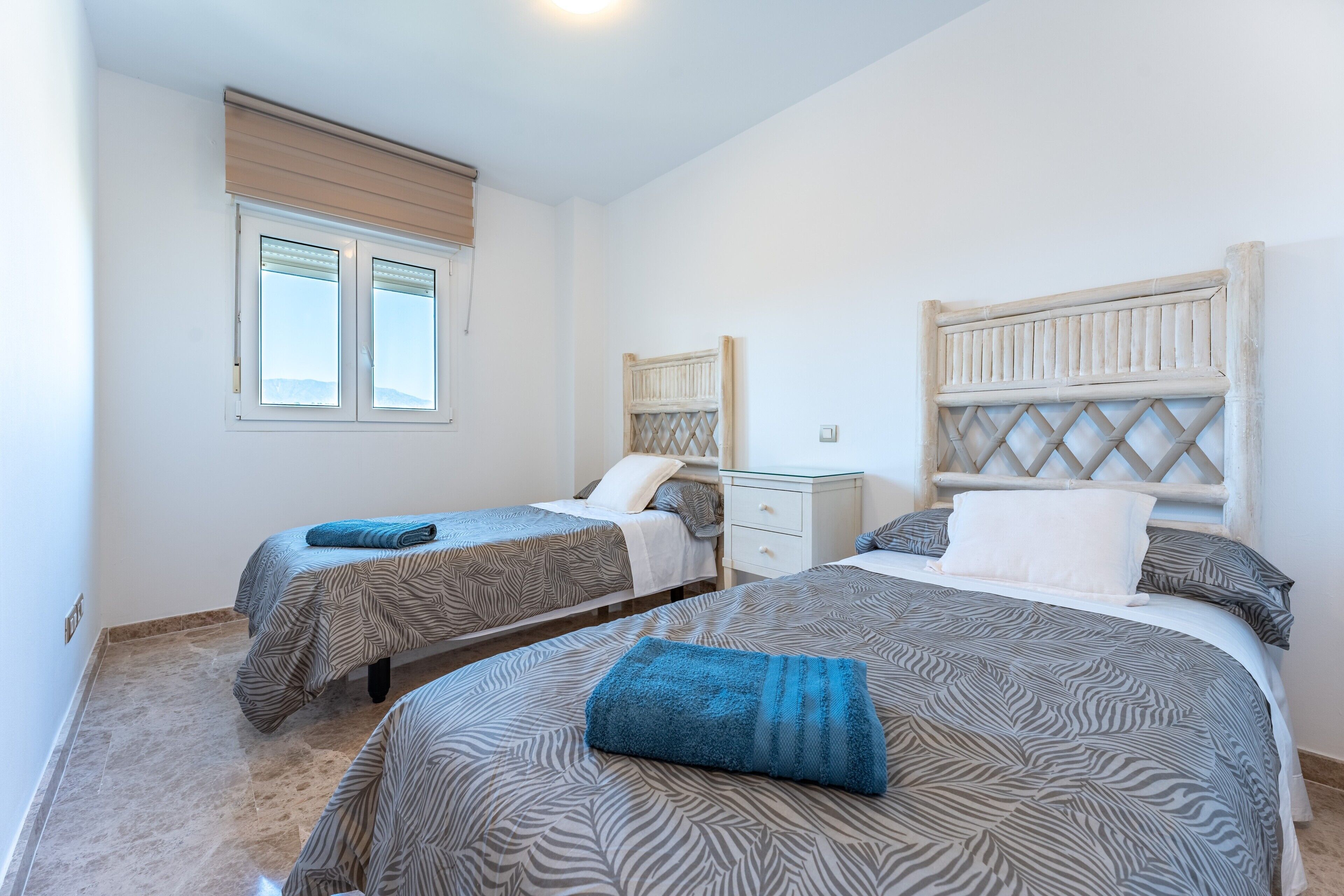 Une chambre avec deux lits, une table de chevet, une fenêtre avec des stores et une tête de lit ornée d’un motif en treillis.