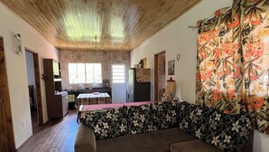 Un salon chaleureux avec un plafond en bois, un canapé agrémenté de coussins à motifs floraux, un tapis rouge et beige, et une cuisine en arrière-plan.