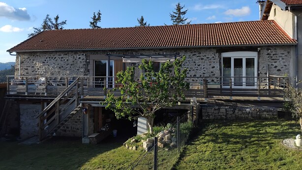 A stone building with a tiled roof, a wooden staircase, and a fenced outdoor area with a small tree.