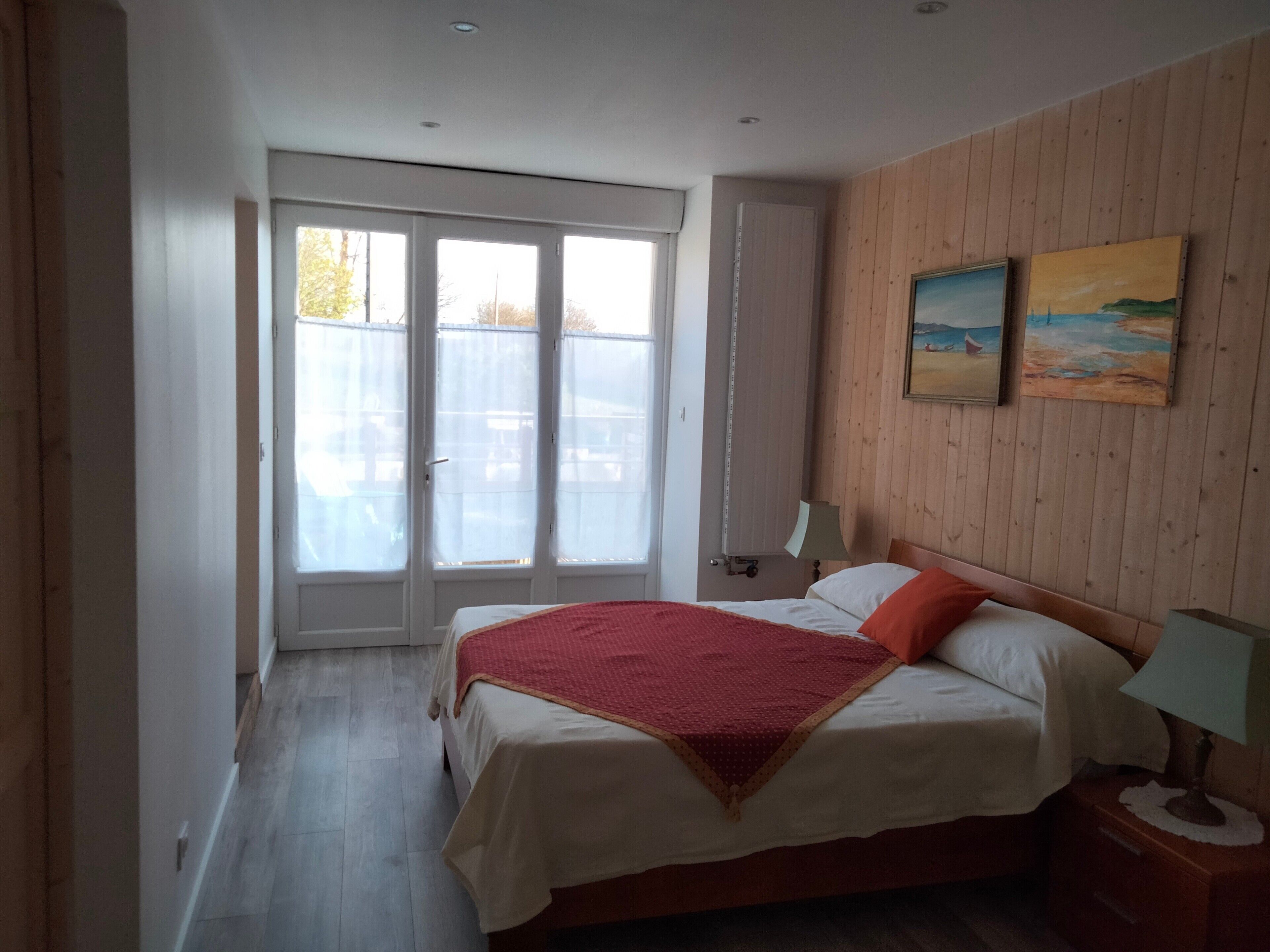 A bedroom with a bed, a nightstand, a lamp, a window with blinds, and a painting on the wall.