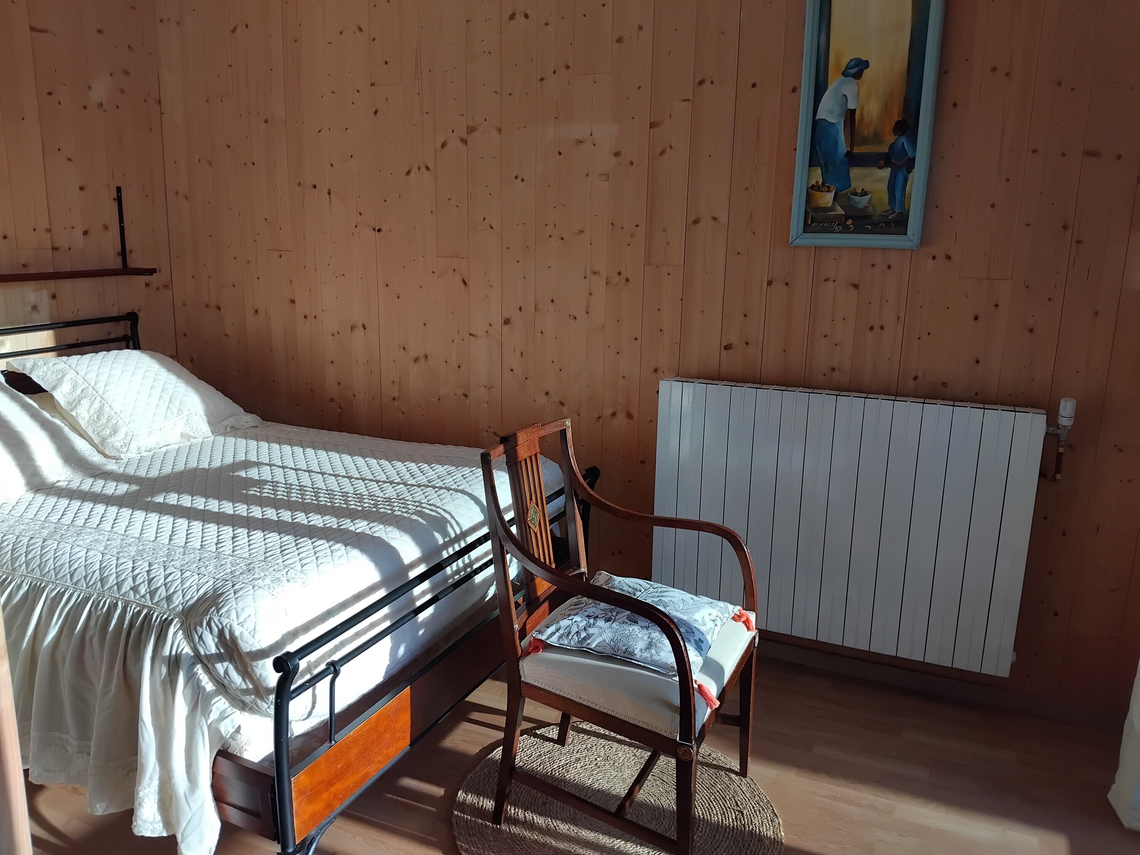 A bedroom with a bed, a chair, a radiator, and a painting on the wall.