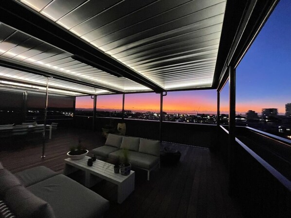 A modern rooftop terrace with a city view at dusk, featuring a sofa, coffee table, and ambient lighting.