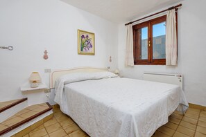 A bedroom with a bed, a window with curtains, a radiator, a wall-mounted lamp, and a small shelf with a lamp. - Holiday Home 'Villetta Palma' with Shared Pool, Private Terrace and Private Garden (Baja Sardinia)