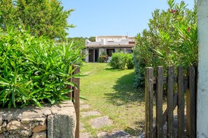 Uma trilha leva a uma casa com telhado de cerâmica, cercada por vegetação e um muro de madeira.