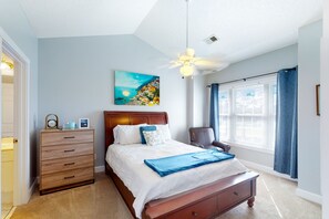 A bedroom with a bed, a dresser, a chair, a ceiling fan, a window with curtains, and a painting on the wall.