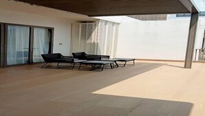 A covered outdoor area with a modern patio, including a seating arrangement and a sliding glass door leading to a balcony. - By The Sea (George Town)