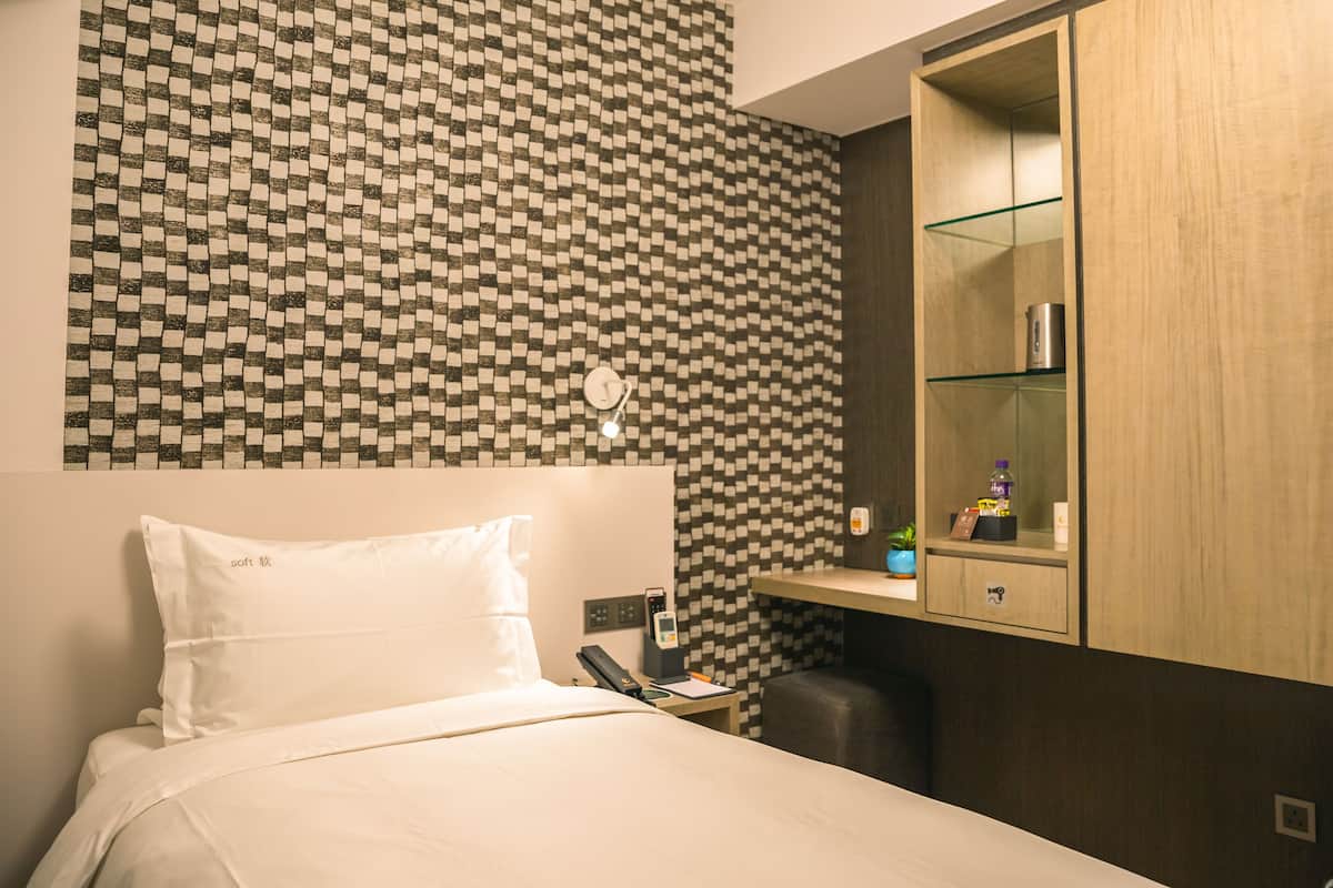 A hotel room with a checkered wall pattern, a bed with white linens, a bedside table with a phone and a lamp, and a wooden cabinet with a mirror and shelves.