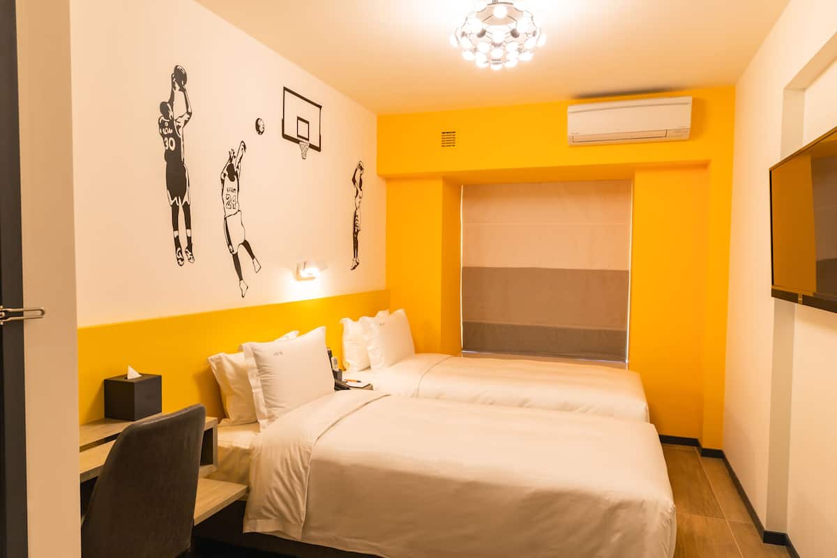 A hotel room with two beds, a desk, a chair, a basketball-themed wall art, and a large window with blinds.
