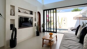 A spacious living room with a ceiling fan, a flat-screen TV, a wooden coffee table, and a sofa with cushions. There are potted plants and a view of the outdoors through large windows. - Villa Cactus Bingin (Pecatu)