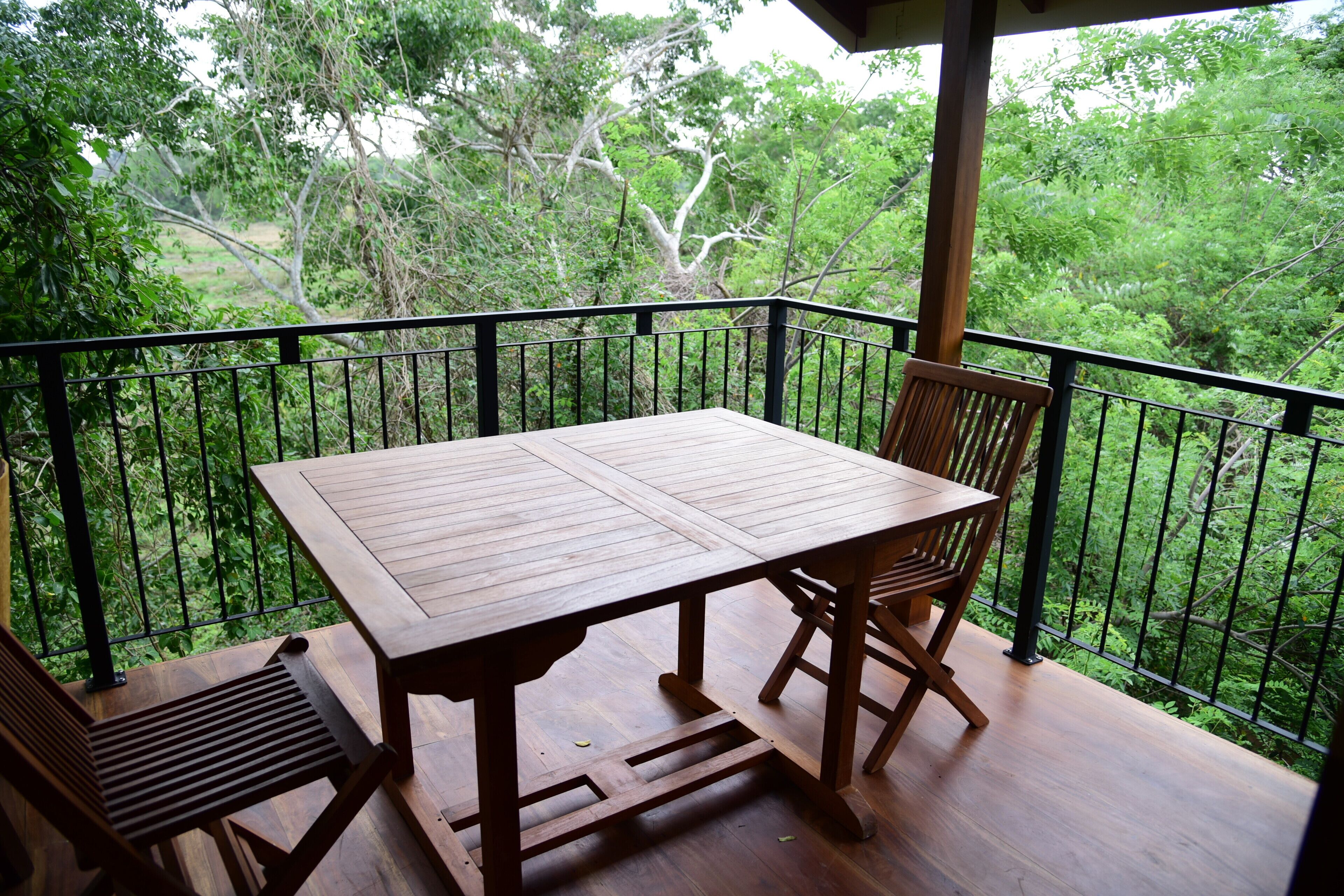 Une table et des chaises en bois sur un balcon doté d’une rambarde en métal, offrant une vue sur un paysage verdoyant et luxuriant.