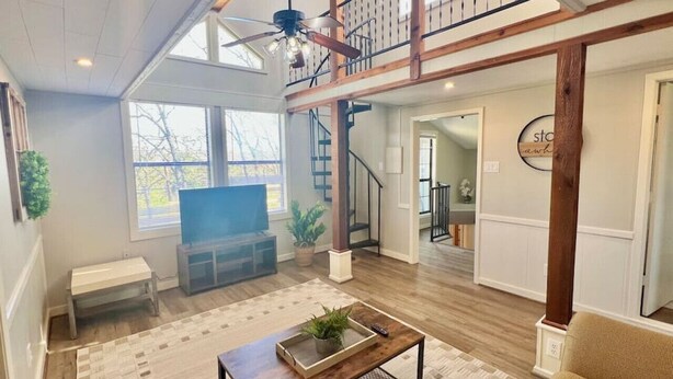 Amplia sala de estar con piso de madera, un ventanal grande, ventilador de techo, escalera y vistas al exterior.