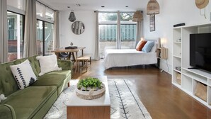 A modern living room with a green sofa, a coffee table with a plant, a bed with pillows, a TV, and a dining area with a table and chairs.