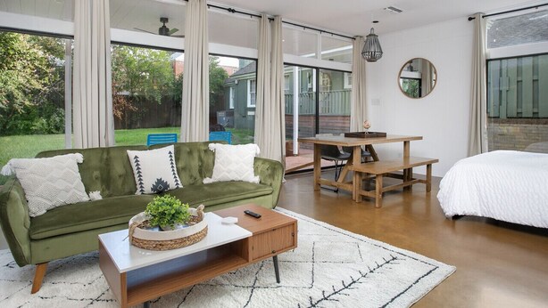A modern living room with a green sofa, a wooden coffee table, a white bed, and large windows offering a view of the backyard.