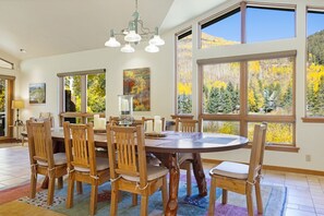 Un espace repas avec une table ronde, des chaises en bois et de grandes fenêtres offrant une vue sur le feuillage automnal.