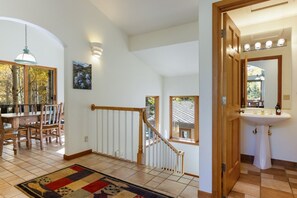 Un comedor con una mesa y sillas de madera, una escalera con barandilla y un baño con lavamanos y espejo.
