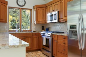 Una cocina con armarios de madera, un refrigerador de acero inoxidable, un microondas, un horno y un ventanal con vista a árboles.