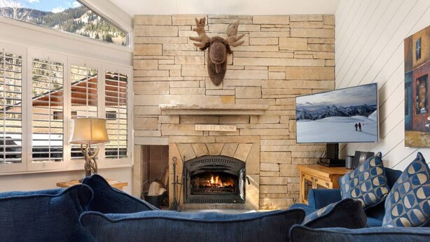 Un salon chaleureux avec une cheminée en pierre, une tête de cerf accrochée au mur et une grande fenêtre équipée de stores.