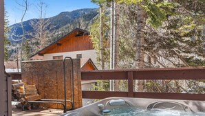 Un bain à remous avec une terrasse en bois, un chalet en bois en arrière-plan et un paysage montagneux.