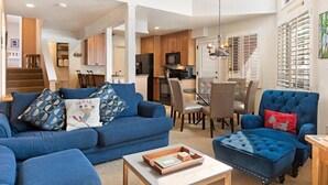 Un salon spacieux avec un canapé bleu, un pouf assorti et un coin repas comprenant une table et des chaises en verre.