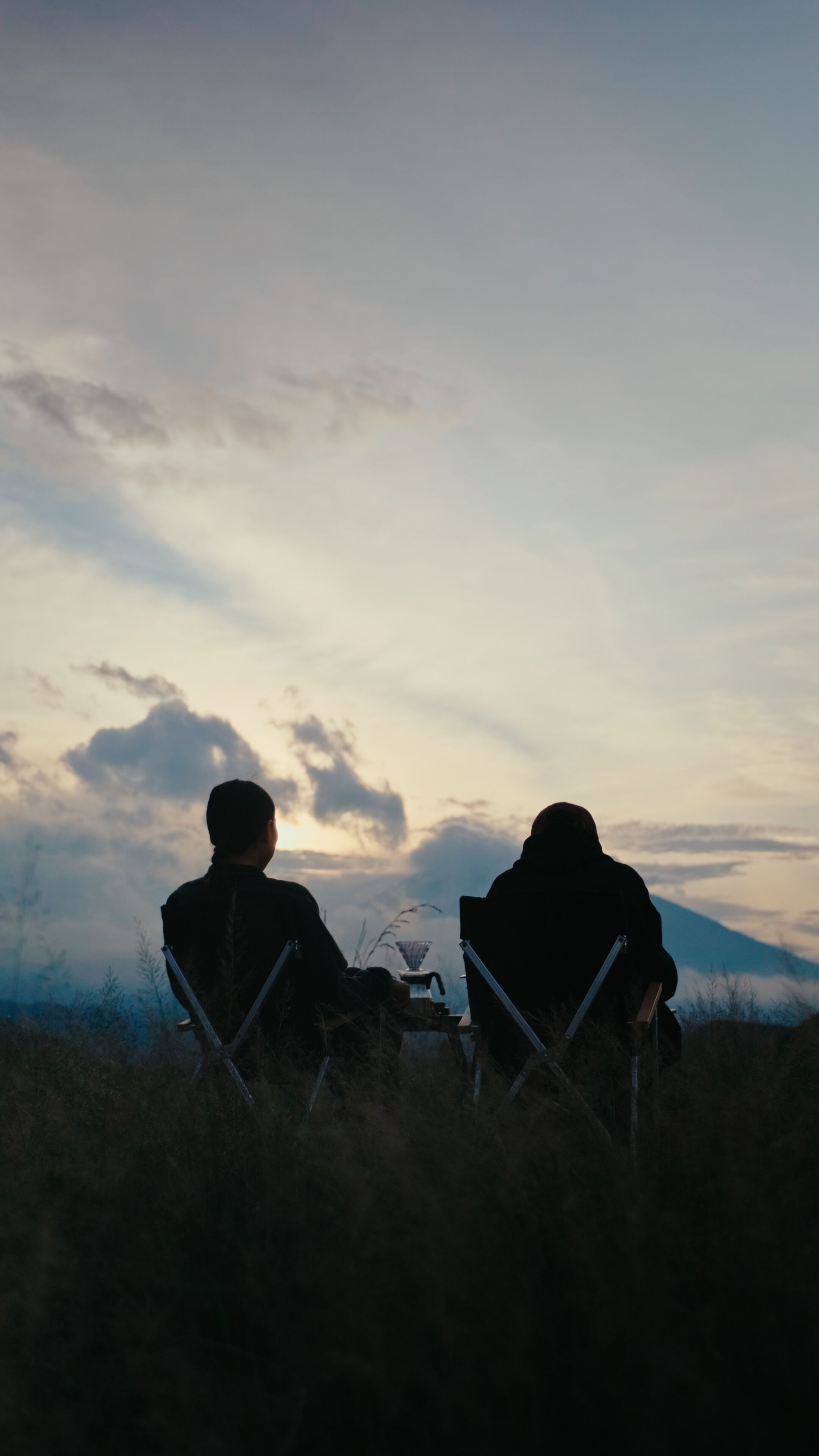 Dos personas sentadas en sillas mirando la puesta de sol con una montaña al fondo.