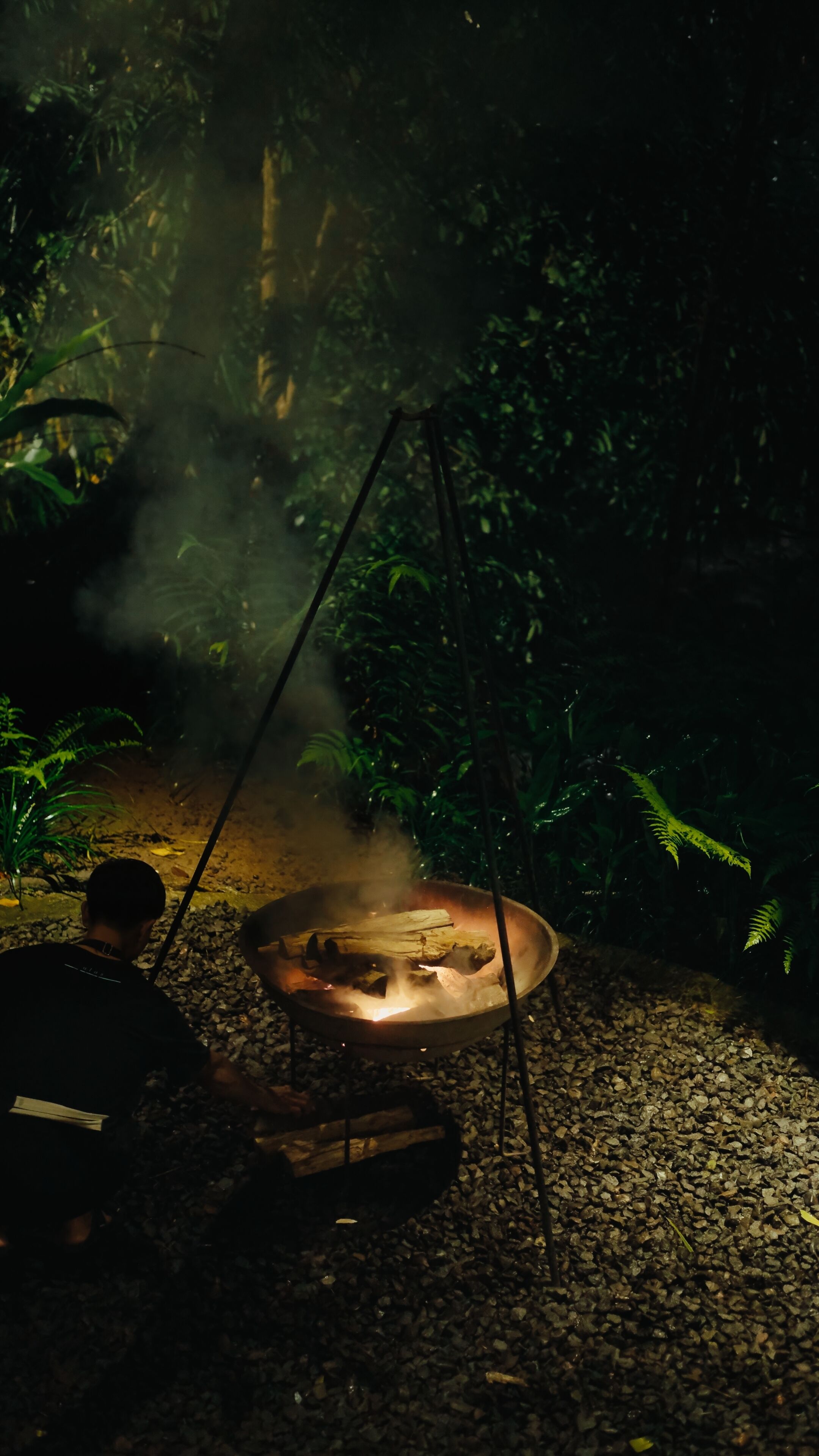 A campfire with logs burning in a metal pan, surrounded by gravel and greenery.