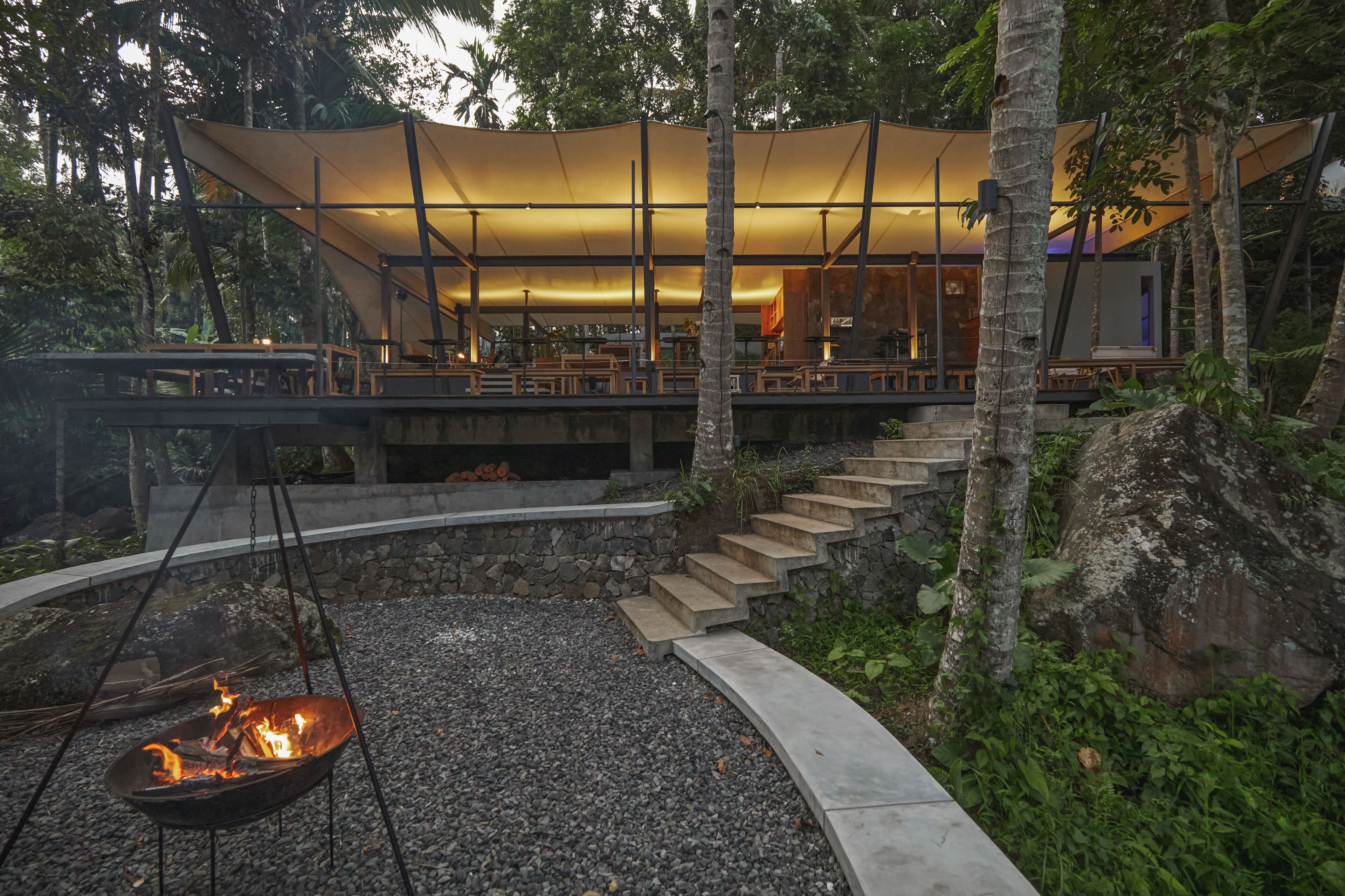 A modern outdoor lounge area with a fire pit, stone steps, and a covered seating area with tables and chairs.