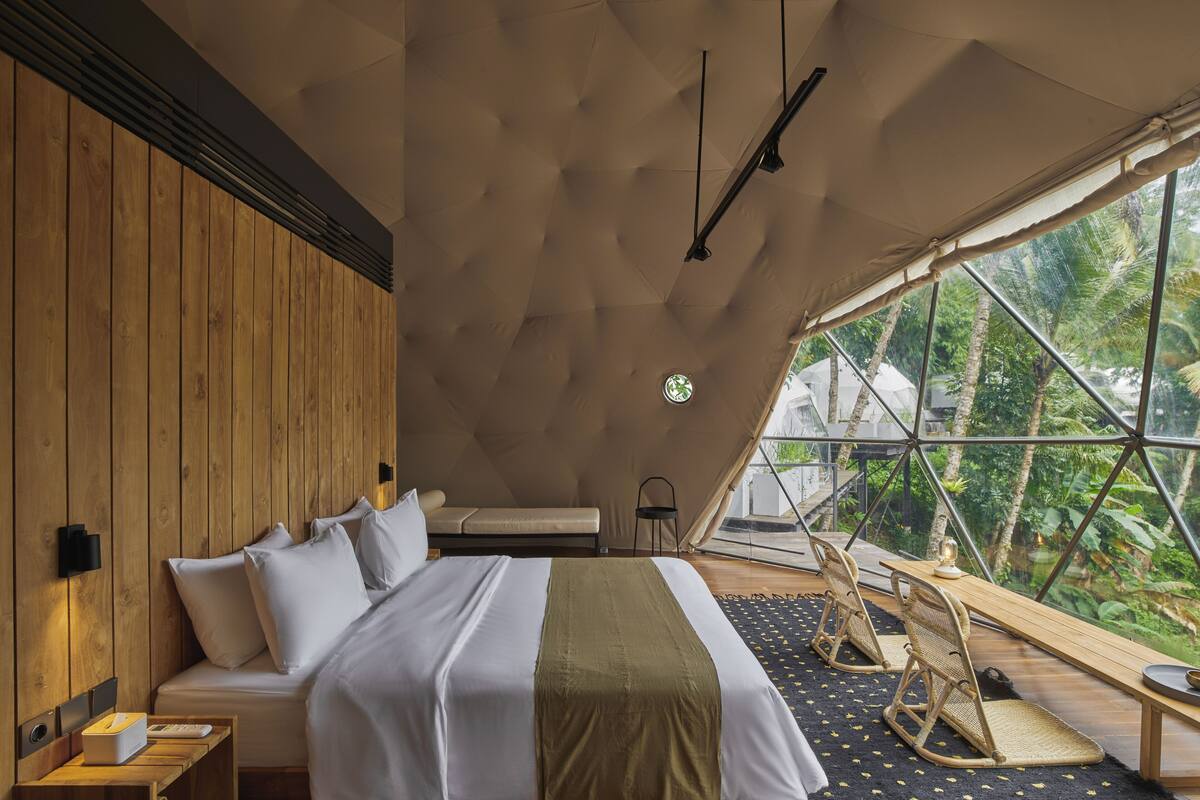 Une chambre aménagée comme une tente, avec un lit, des murs recouverts de panneaux de bois, une vue sur la verdure et une petite table avec des chaises.