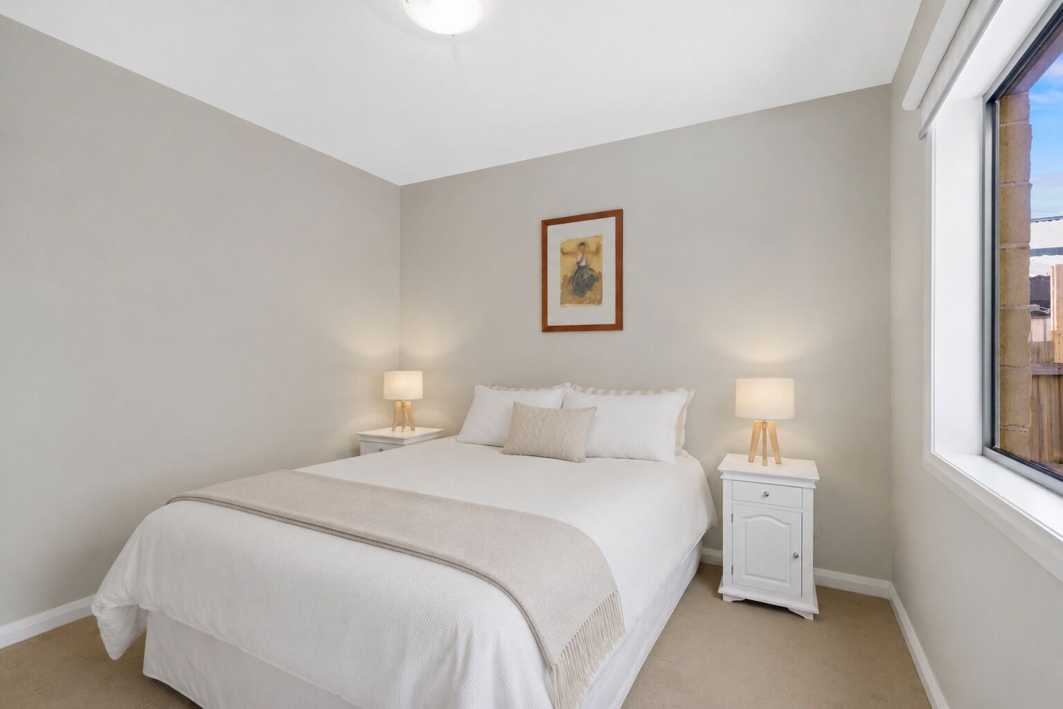 A bedroom with a bed, two bedside tables, a window, and a wall-mounted picture.