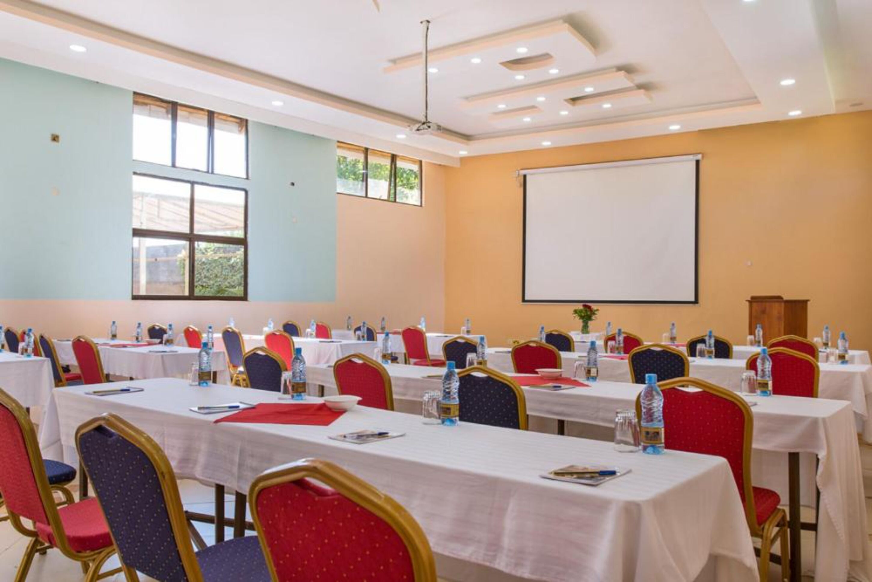 A conference room with multiple tables and chairs, a projector screen, and windows with natural light.