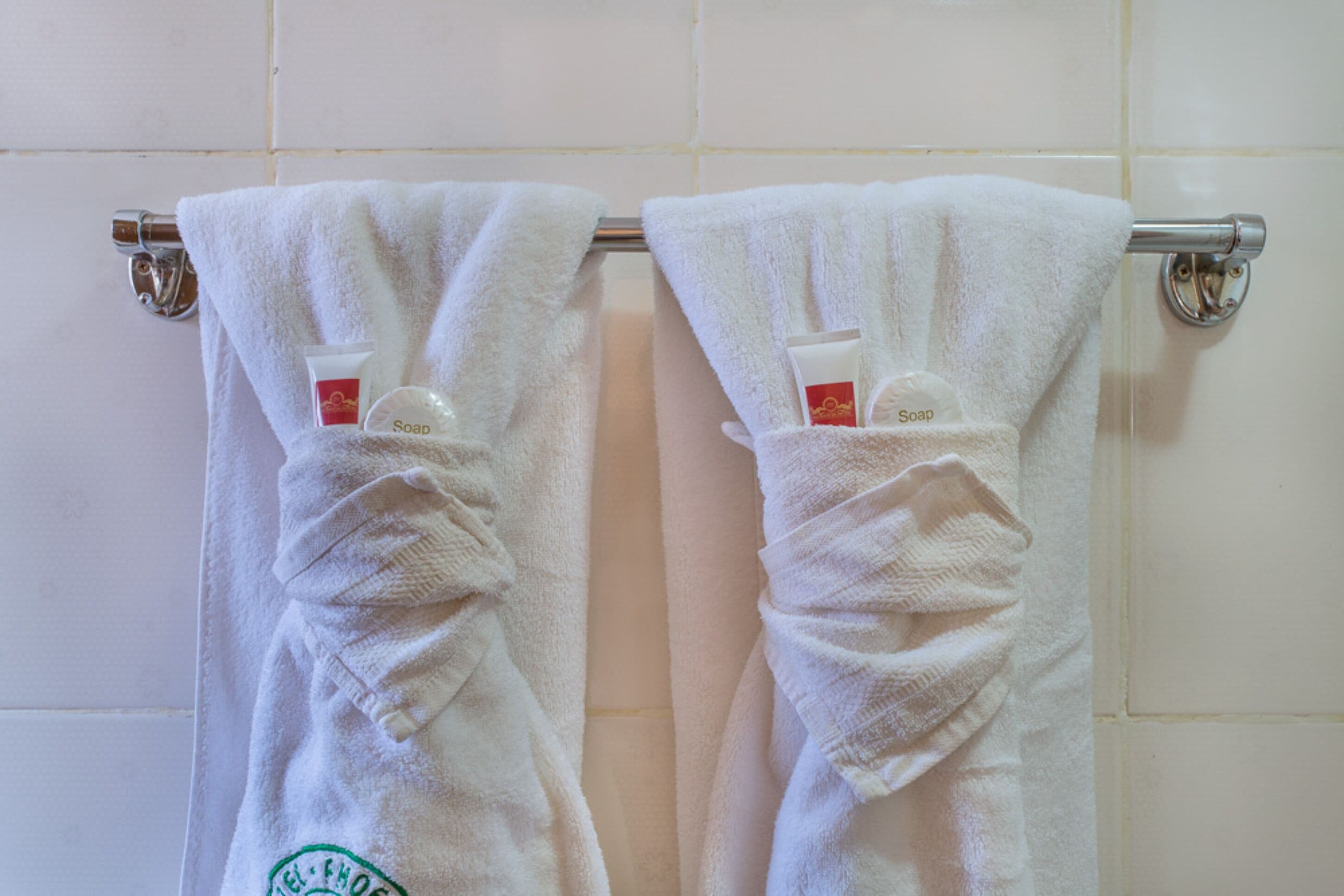 Deux serviettes blanches sont suspendues à un porte-serviettes en métal dans une salle de bain dont les murs sont recouverts de carreaux blancs.