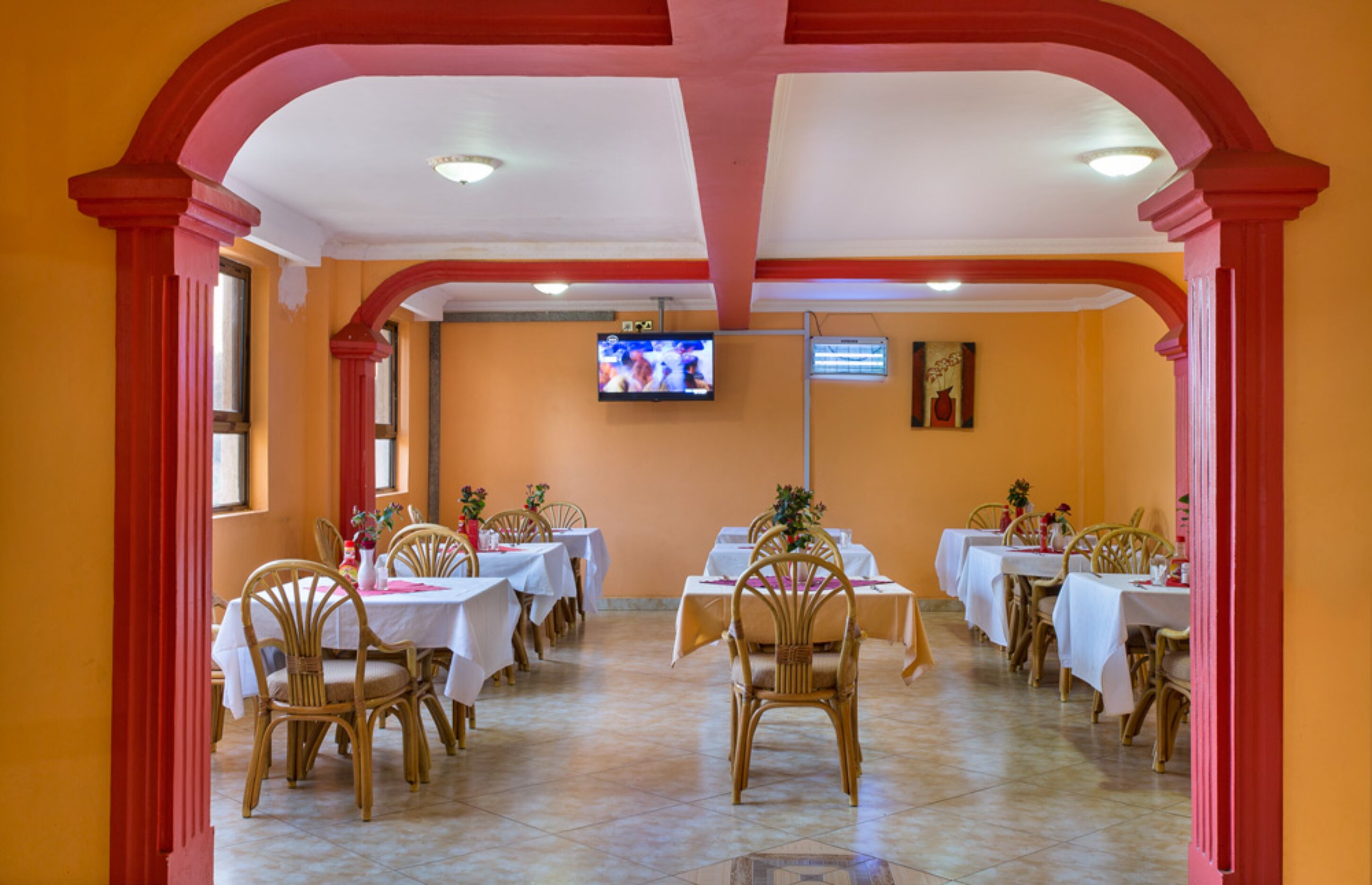 Uma área de refeições com mesas e cadeiras, uma televisão presa à parede e piso revestido de azulejos.