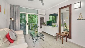 Ein Wohnzimmer mit Sofa, Couchtisch und Fernseher. Ein Balkon bietet Blick auf Grünflächen.