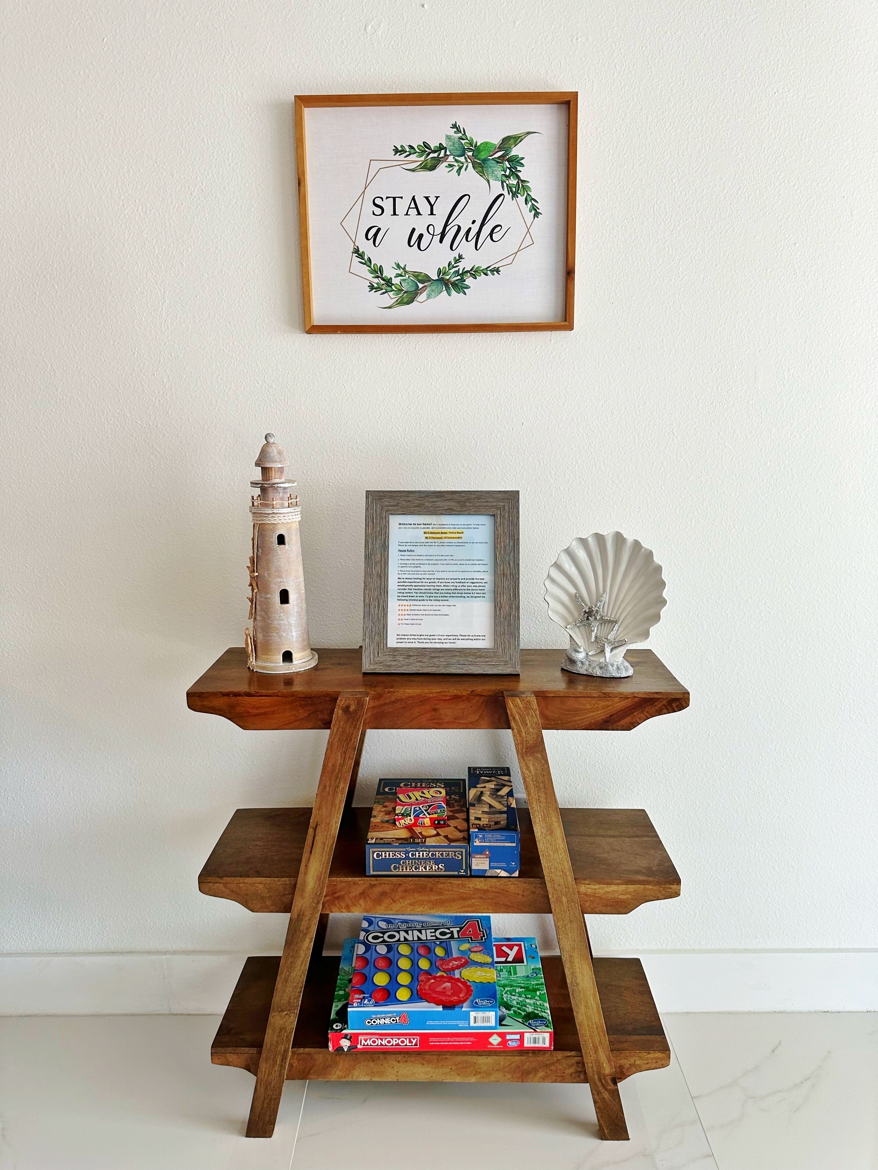 Ein Holzregal mit einem gerahmten Schild mit der Aufschrift "STAY a while", einem Leuchtturmmodell, einem gerahmten Willkommensschild und Gesellschaftsspielen.