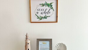 A wooden shelf with a framed sign saying "STAY a while", a lighthouse model, a framed welcome sign, and board games.