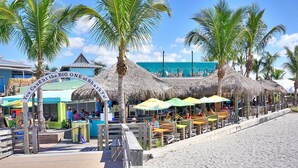 A beachside restaurant with thatched umbrellas, a wooden deck, and palm trees.
