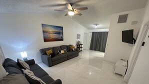 A living room with a sofa, a ceiling fan, a painting, and a television.