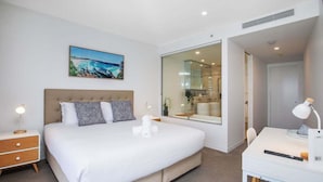 Quarto moderno com cama grande, mesas de cabeceira, escrivaninha e banheiro visível através de uma porta de vidro.