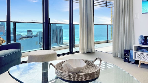 Uma sala de estar moderna com uma mesa de vidro, uma cadeira de fibra natural e vista para o oceano e a paisagem urbana.