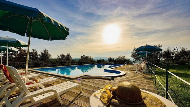 Ein Poolbereich mit Liegestühlen und Sonnenschirmen, ein Hut auf einem Tisch und Blick auf den Sonnenuntergang über dem Wasser.