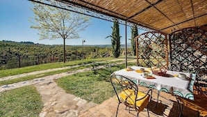 Un área de comedor al aire libre con una mesa preparada para dos, una pérgola arriba y vistas a un árbol y al paisaje.