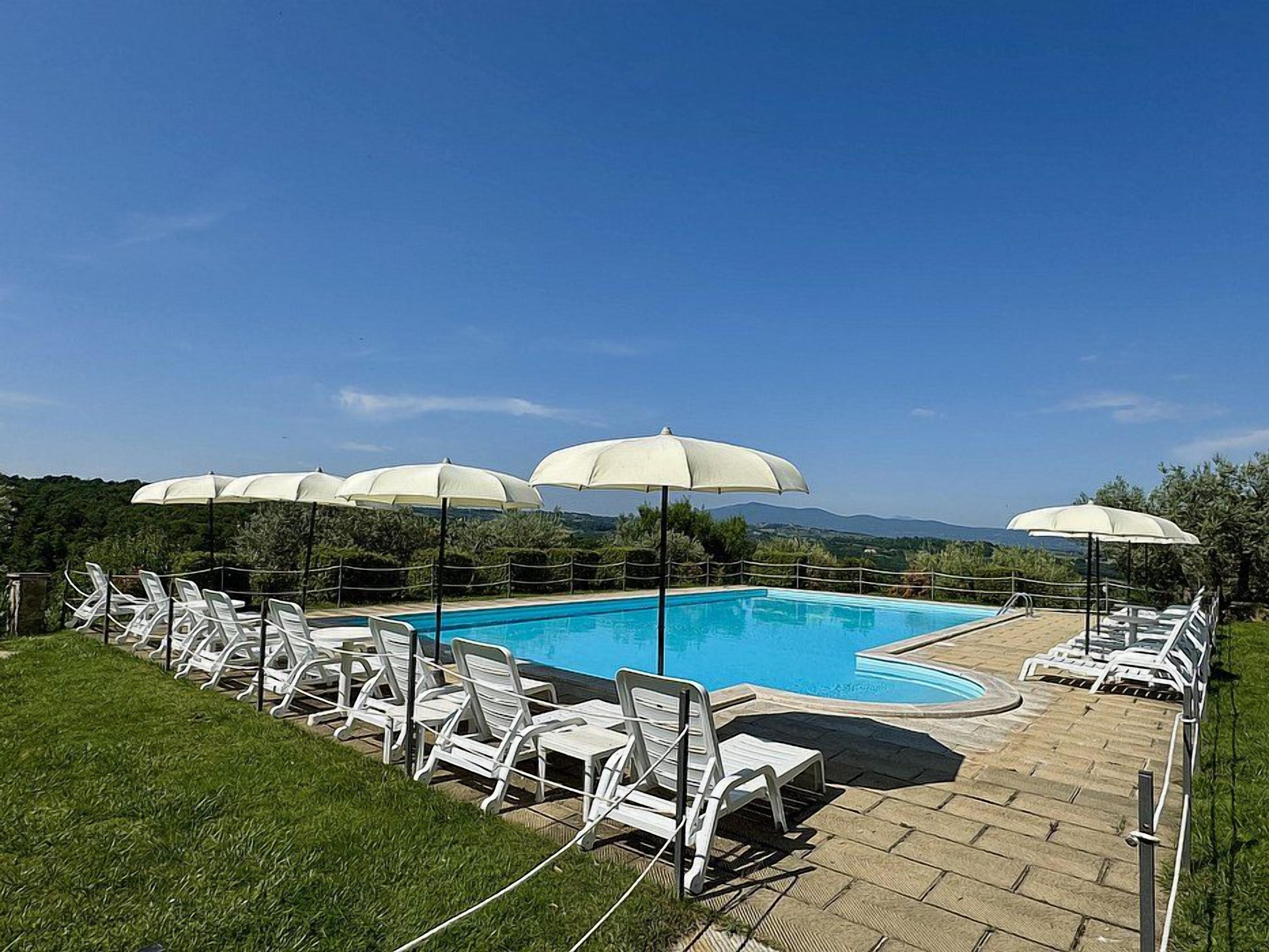 Piscina con sillas de descanso y sombrillas, rodeada por una valla, bajo un cielo despejado y montañas a la distancia.