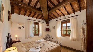 A bedroom with a wooden ceiling, a bed with a patterned bedspread, two windows with curtains, a bedside table with a lamp, and a wall-mounted shelf.