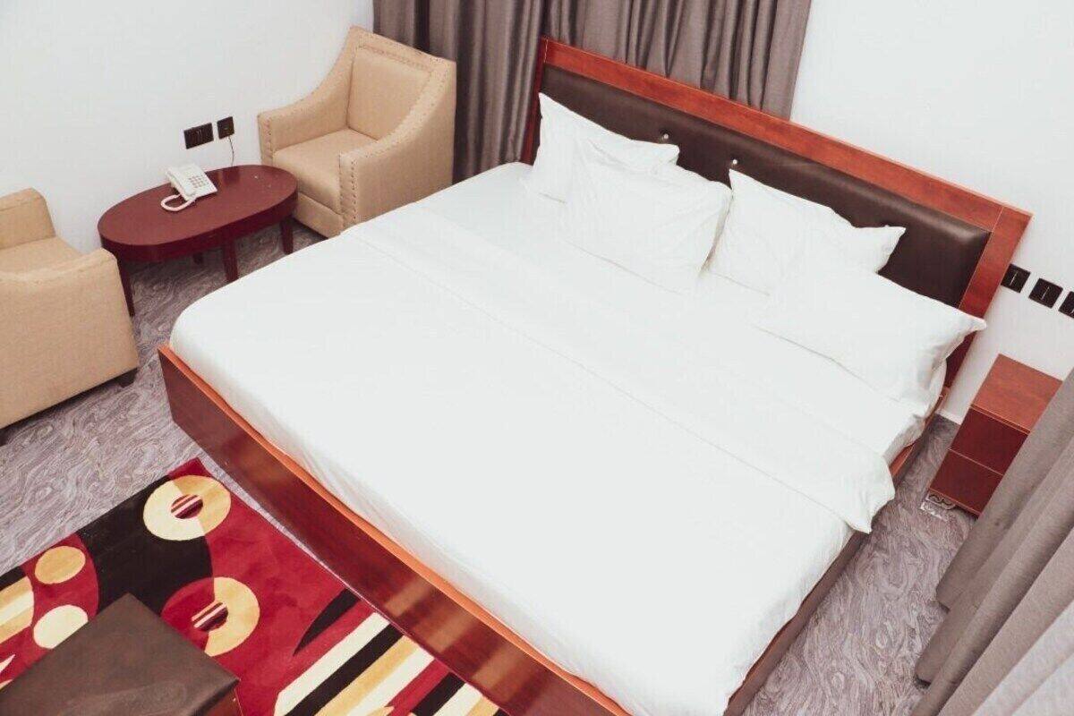 Une chambre d’hôtel avec un grand lit, deux fauteuils, une petite table ronde avec un téléphone et un tapis rouge à motifs.